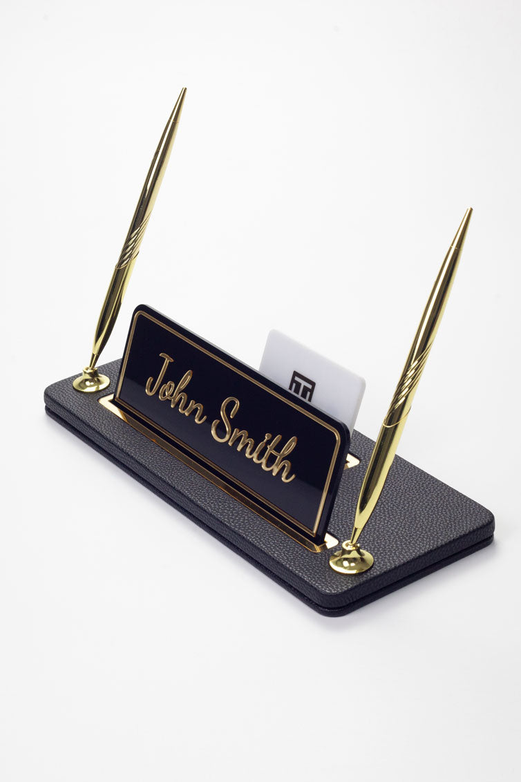 Siyah deri masa isimliği, altın detaylı iki kalem ile, "John Smith" yazılı plaka ve kart tutuculu, yan açıdan görünüm.